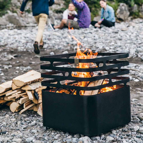 Brasero Cube Hofats noir allumé avec un tas de bûches à proximité