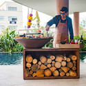 Chef cuisinier en action sur le brasero OFYR Island 100 corten à côté d'une piscine