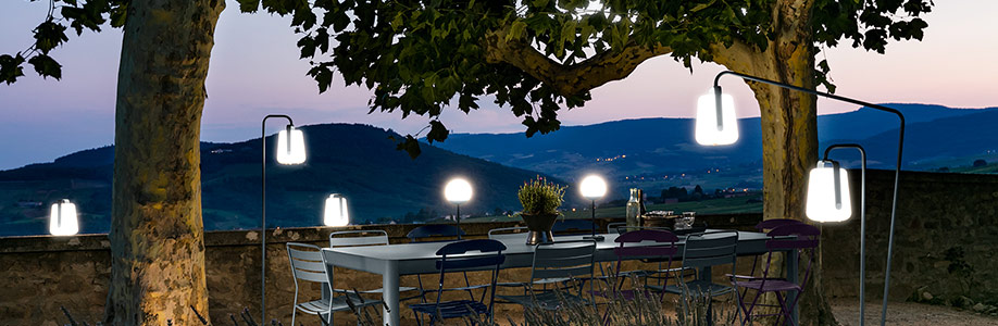 Plusieurs lampes Balad Fermob posées et suspendues sur une table de jardin sous les arbres