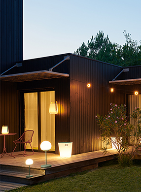Plusieurs lampes Fermob allumées et installées autour d'une maison en bois sur la terrasse extérieure