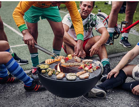 Cuisson sur plancha et grille installé sur le brasero barbecue Bowl Hofats bas entouré par des joueurs de foot