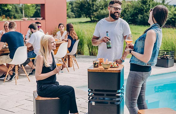 Plusieurs utilisations de la tablette en bambou sur le brasero Cube Höfats noir posé sur une terrasse